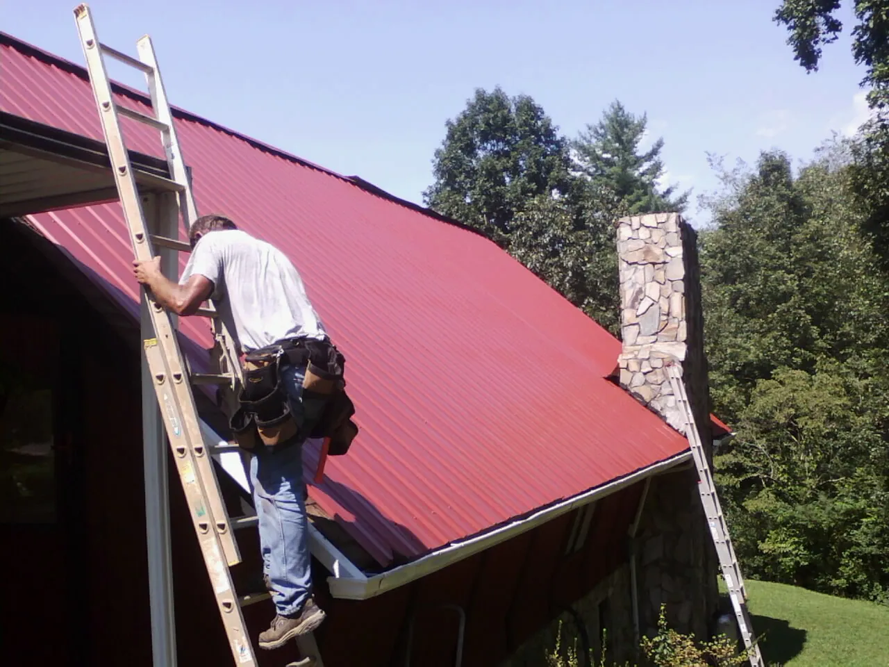 Roofer on a ladder beside a red metal roof and stone chimney — Soffit & Fascia Repair by Roofing Brothers in Verndale
